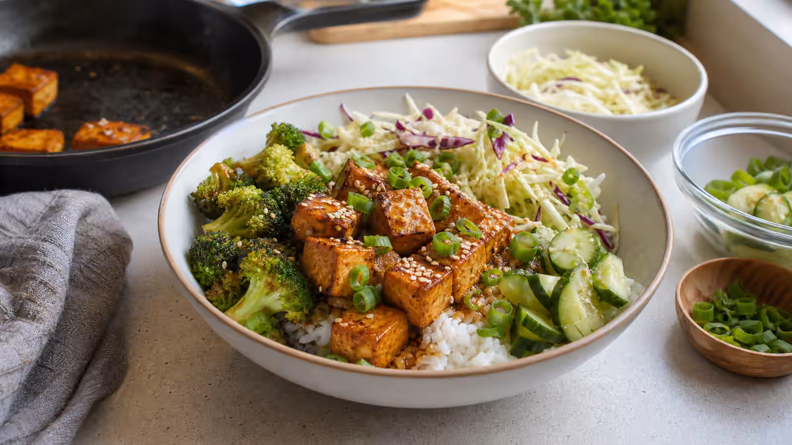 A rice bowl topped with browned tofu cubes, broccoli, slaw, cucumber, green onion, sesame, and savory sauce.
