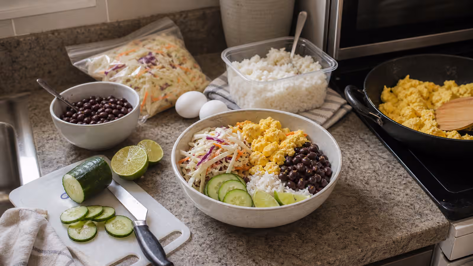 A compact vacation-rental kitchen counter with a rice bowl, small skillet, microwave rice, beans, slaw, cucumber, eggs, and limes.
