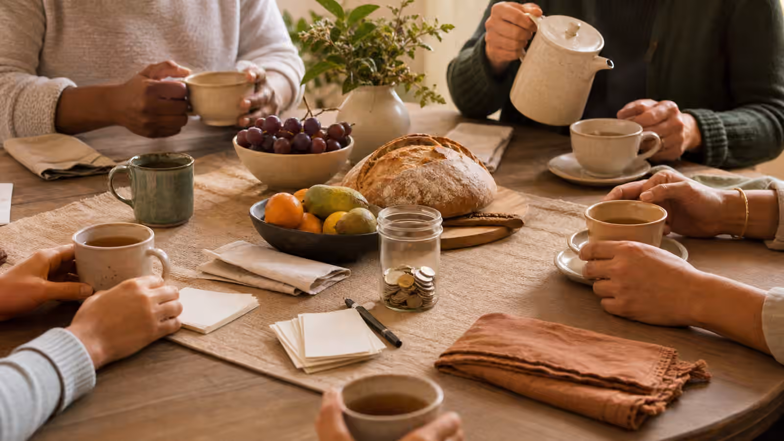 a shared table with bread, fruit, tea, folded napkins, blank cards, and a small jar of optional coins