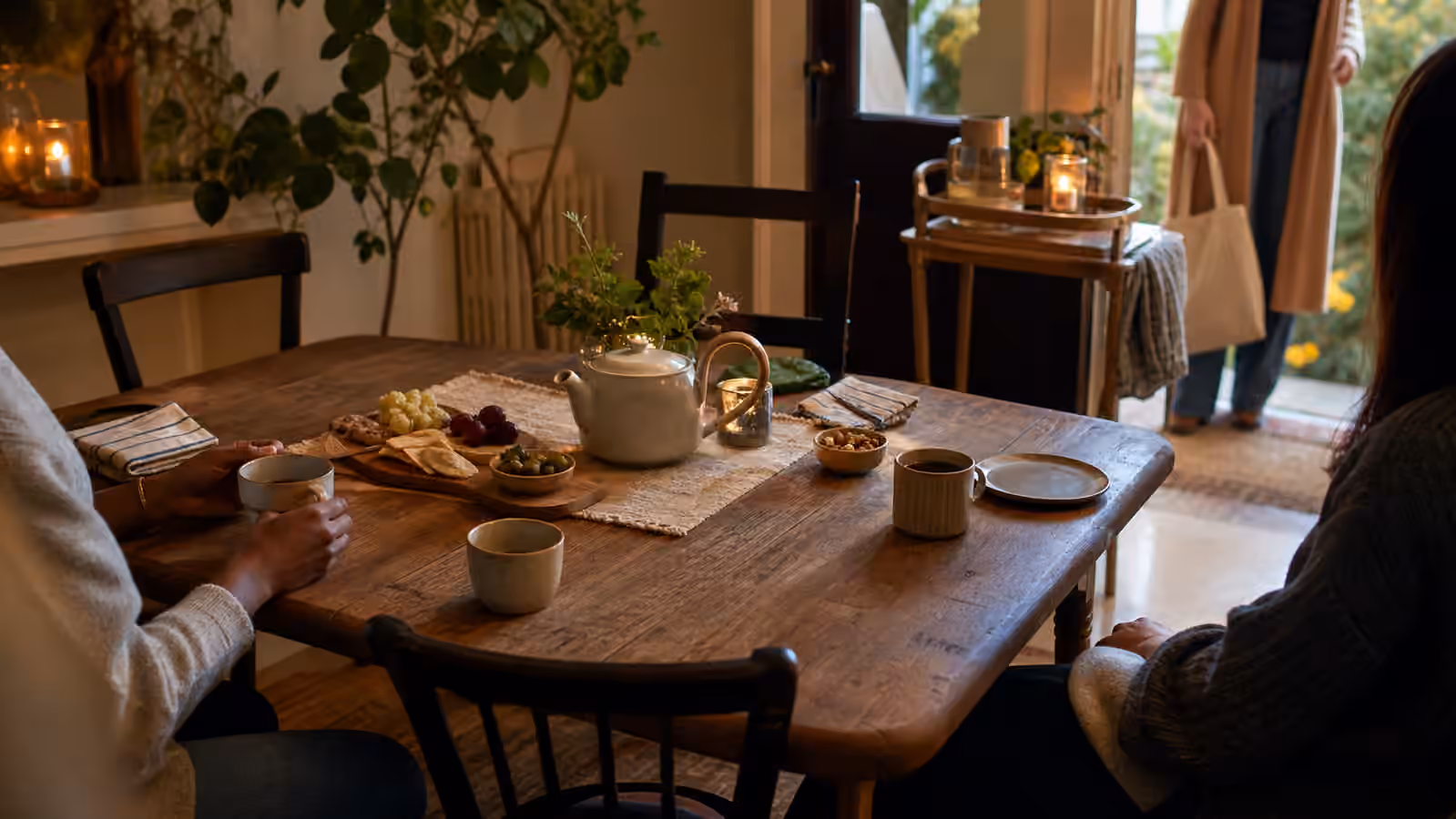 a warm small table with tea, snacks, two seated guests, two empty chairs, and a tray near the door