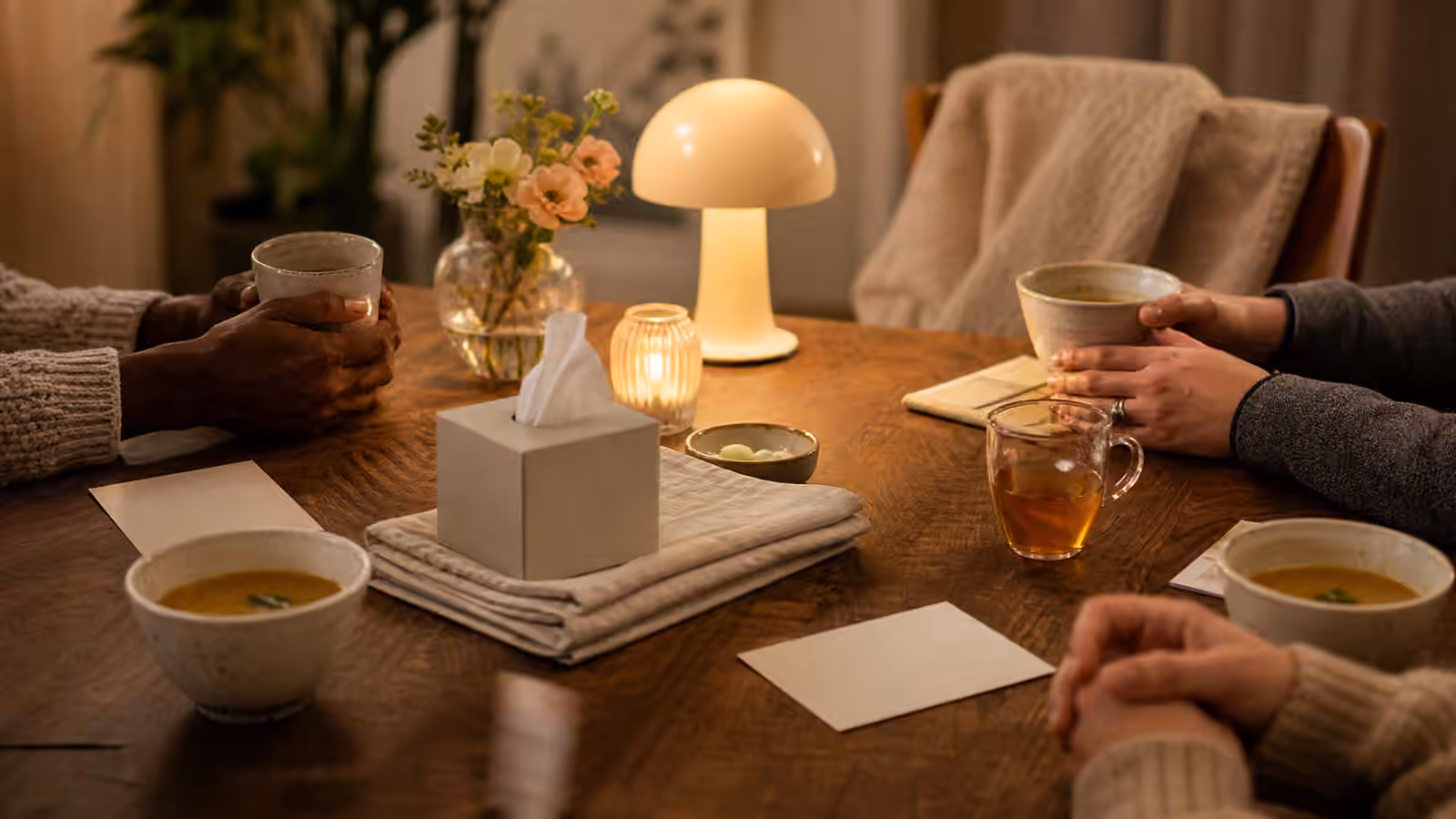 a soft evening table with soup bowls, tea, napkins, discreet tissues, a blanket on one chair, and warm lamplight