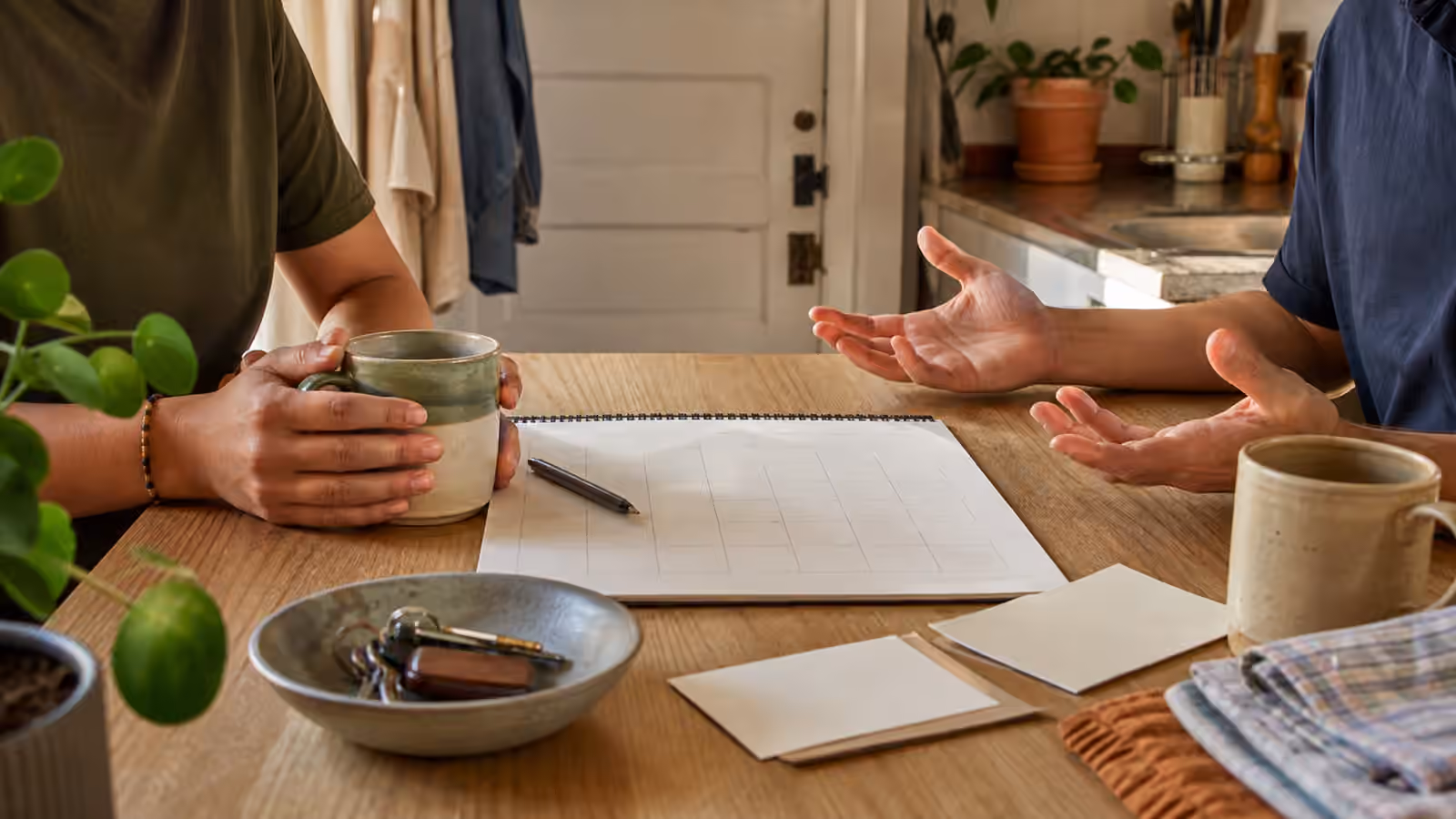 two mugs, keys, blank house notes, folded towels, and a calendar on a shared apartment kitchen table