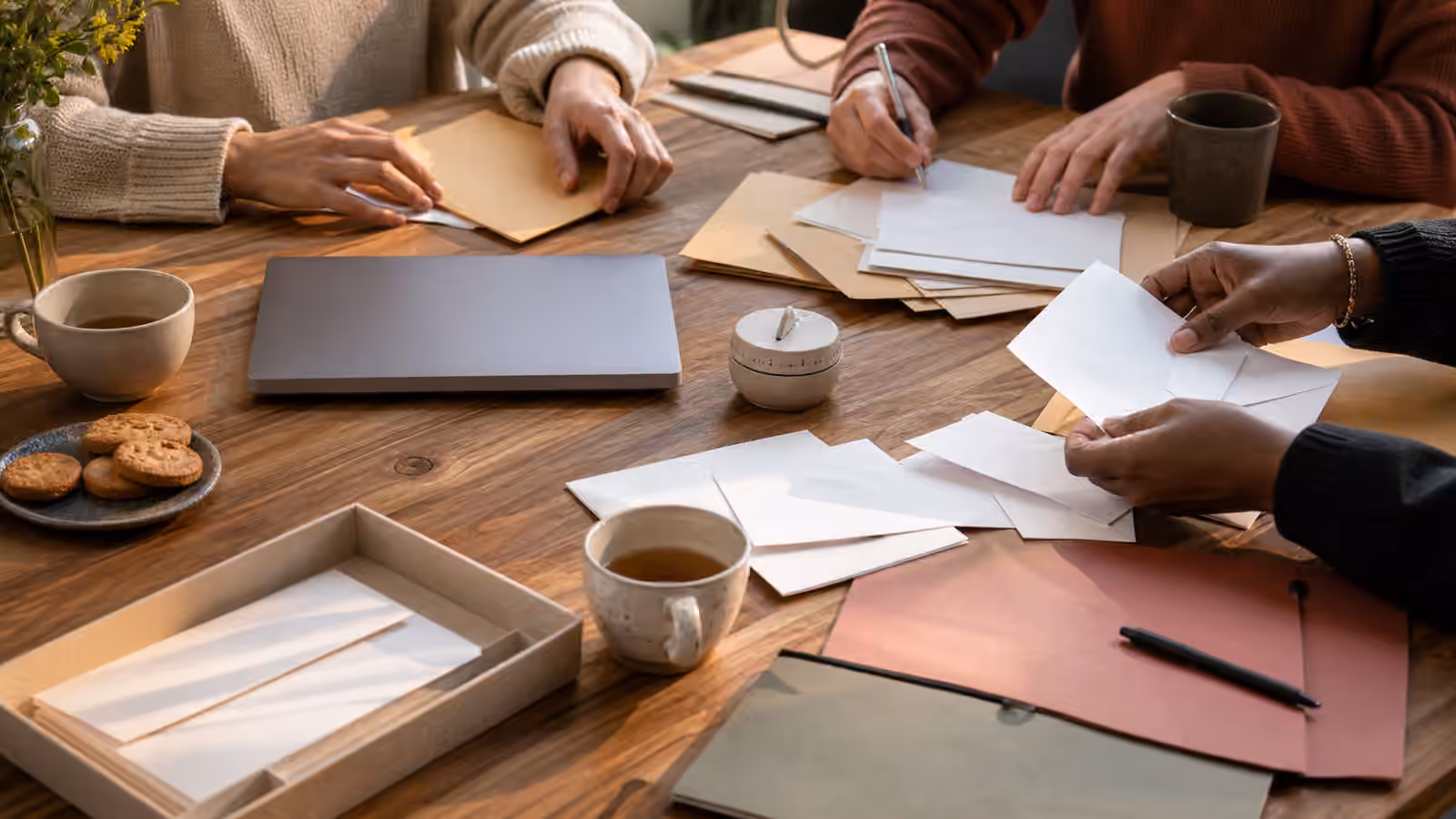a shared table with blank envelopes, folders, pens, tea cups, a closed laptop, simple snacks, and hands sorting papers