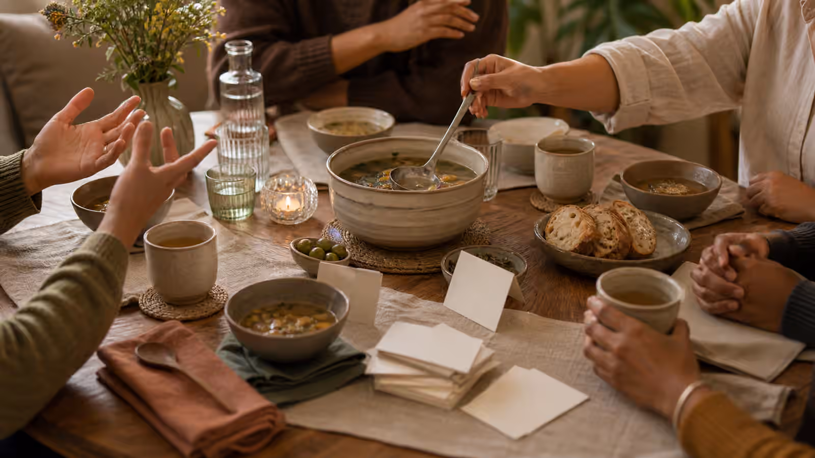 a small soup table with hands, cups, blank conversation cards, and a host moving a serving spoon toward the center