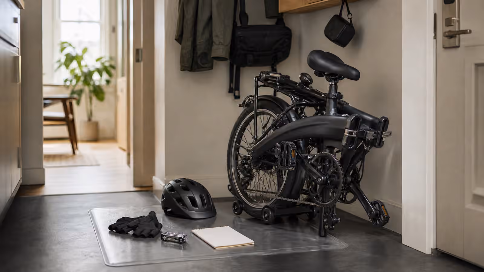 A folding electric bike in a compact hallway with hinge hardware, latch, helmet, gloves, small tool, floor mat, and blank route note.