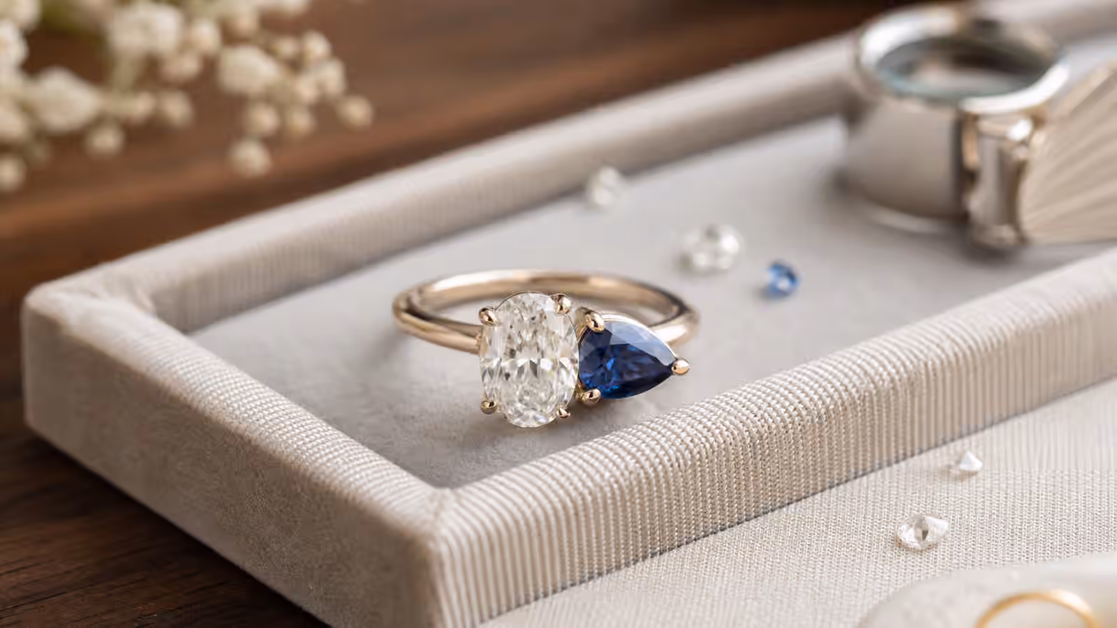 Two-stone engagement ring with an oval diamond and pear-shaped blue sapphire on a linen jeweler's tray.