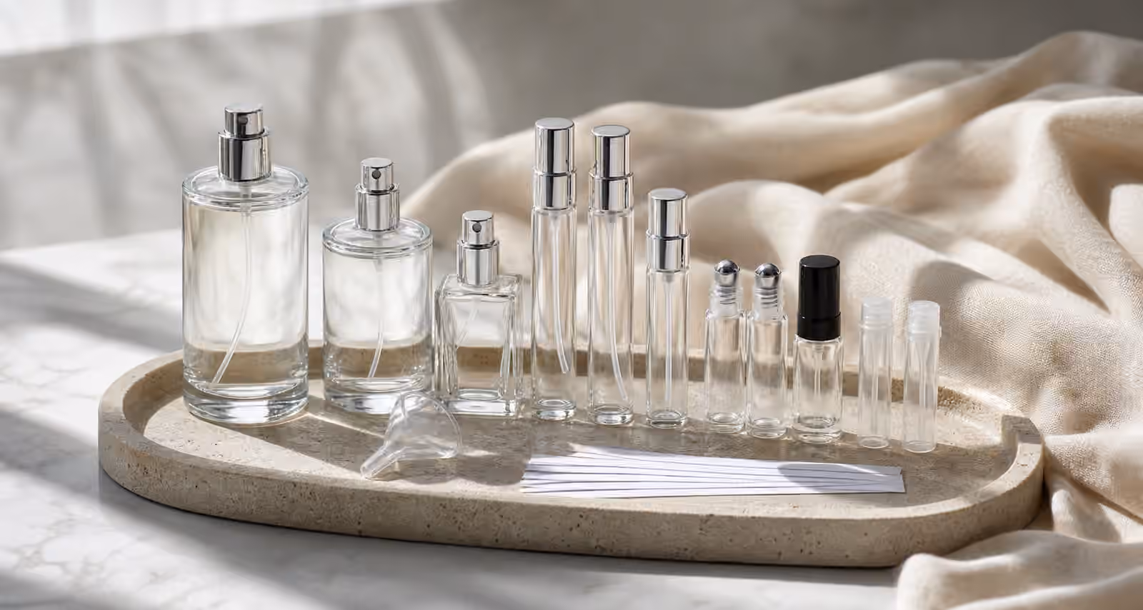 Unbranded perfume atomizers, rollerballs, sample vials, travel sprays, blotter strips, and pale fabric arranged on a fragrance studio tray.