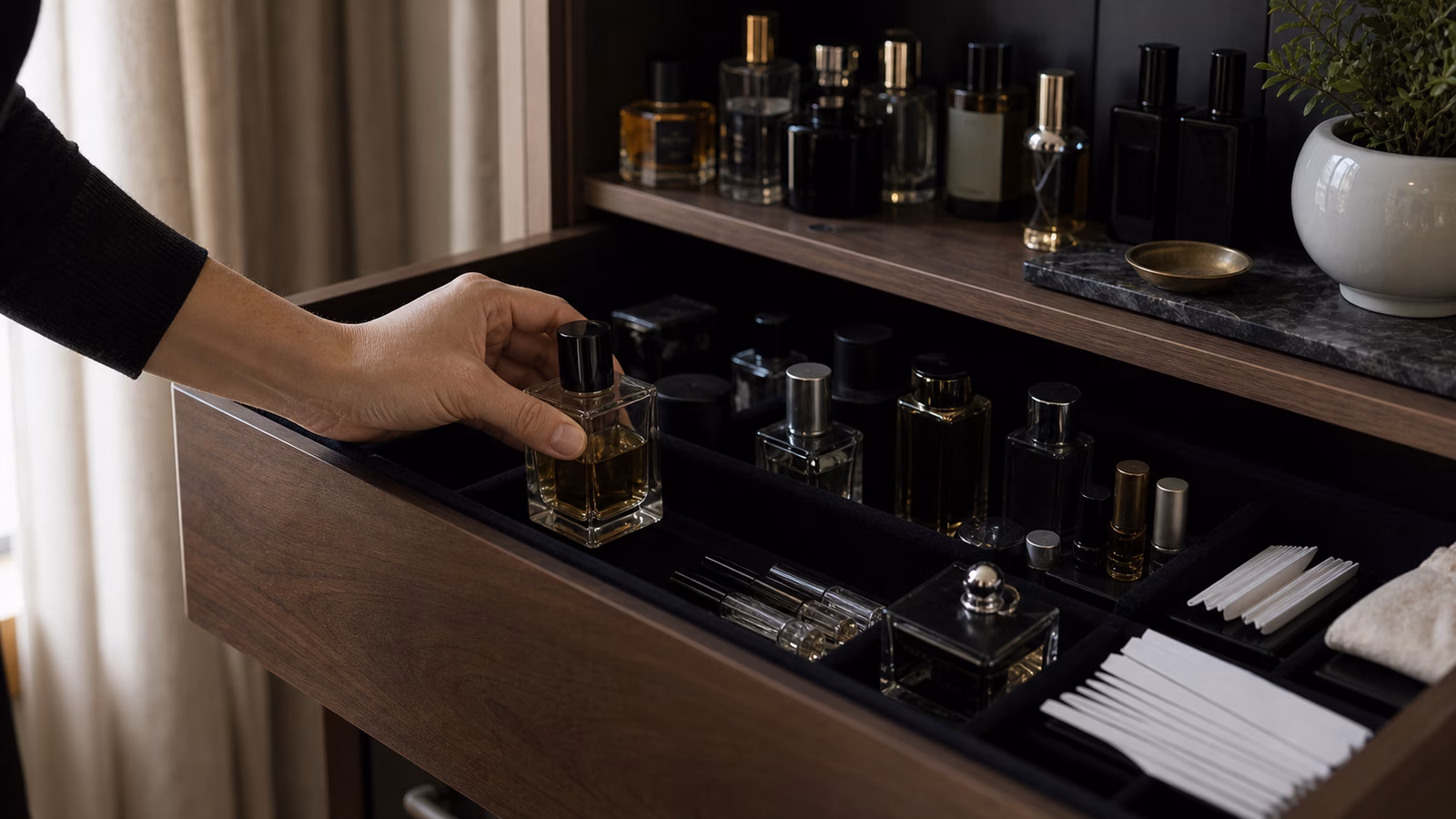 A hand places an unbranded perfume bottle into a dark lined dresser drawer with sample vials, blotters, trays, and shaded fragrance storage.
