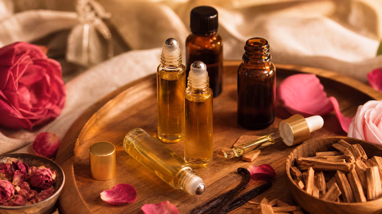 Perfume oil rollerballs, small amber bottles, a carrier oil dropper, vanilla, sandalwood, and rose petals on a warm studio surface.