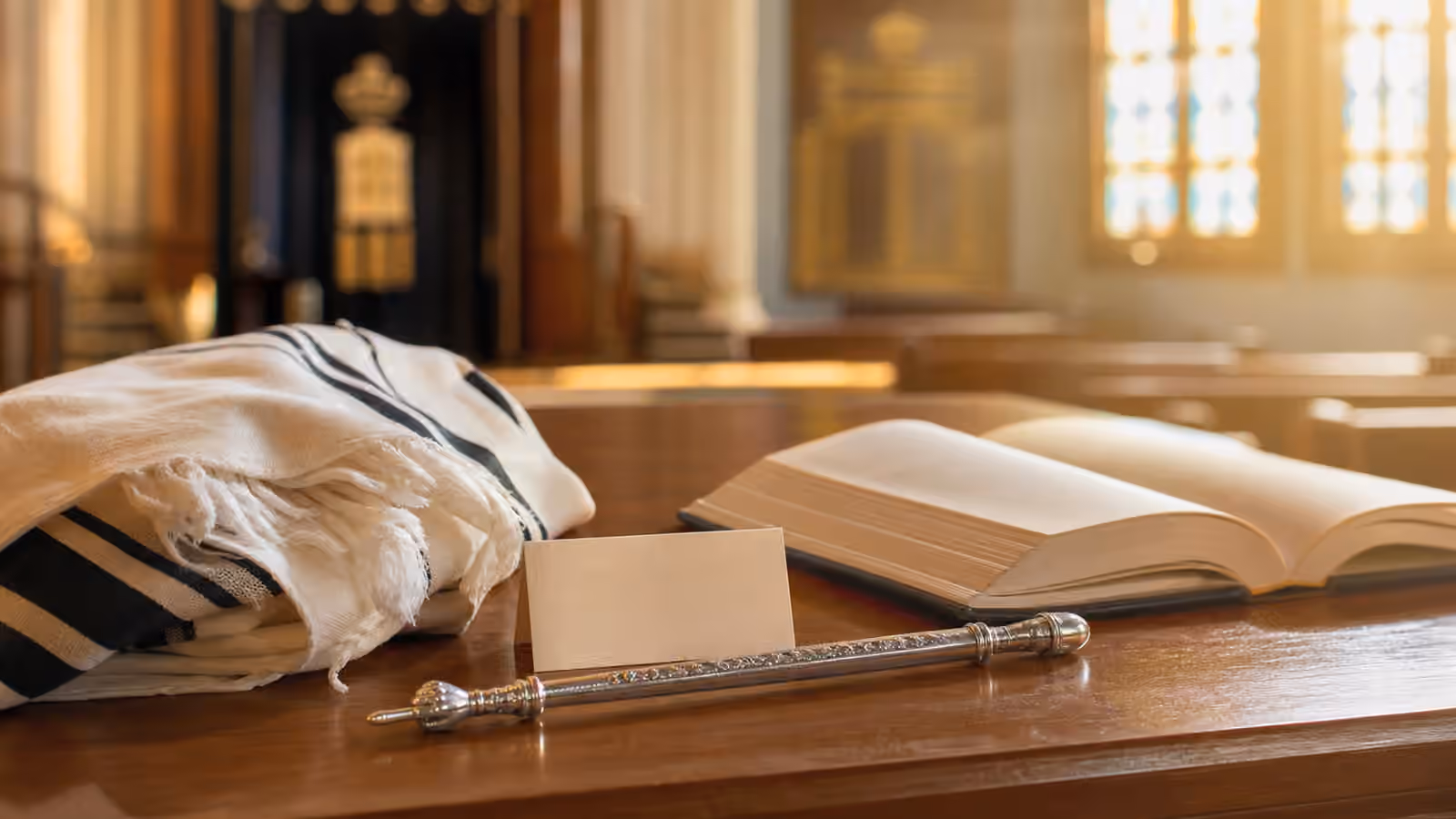 A synagogue reading table with folded tallit, blank honor card, open book, and silver Torah pointer in warm light.