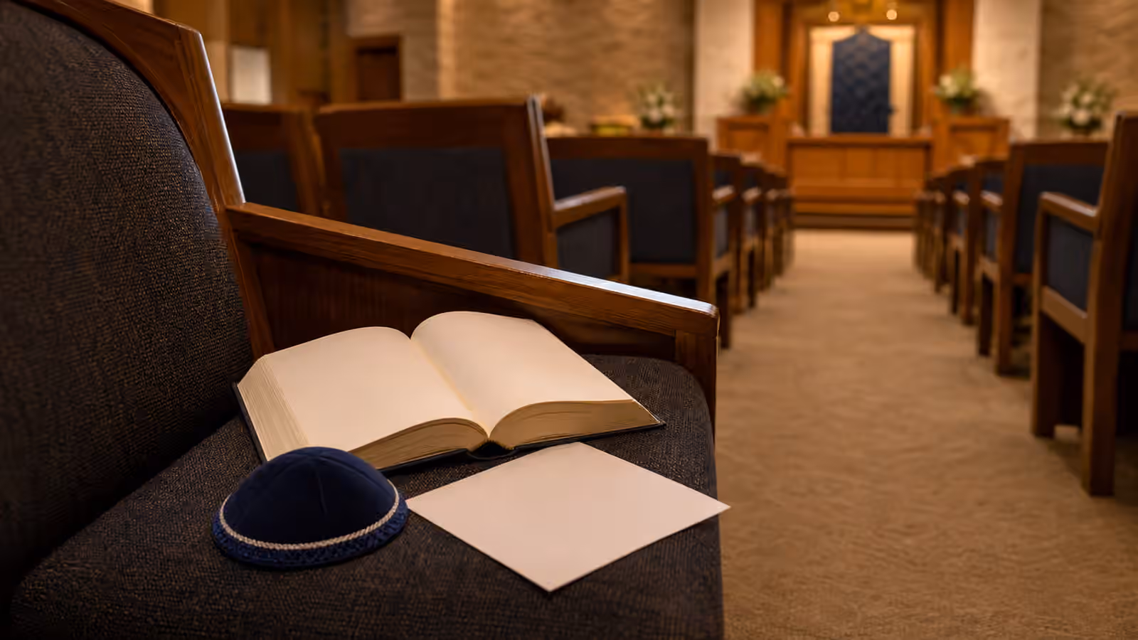 A synagogue seat prepared for Kabbalat Shabbat with an open prayer book, folded kippah, unmarked song sheet, and a quiet sanctuary aisle.
