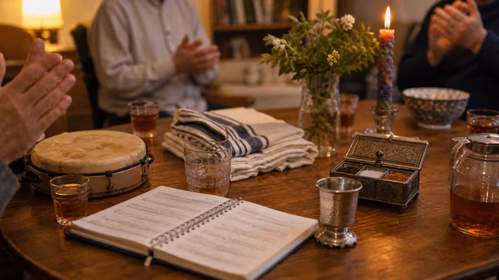 A warm Jewish singing table with songbook, drum, spice box, candle, cup, tea, and clapping hands.