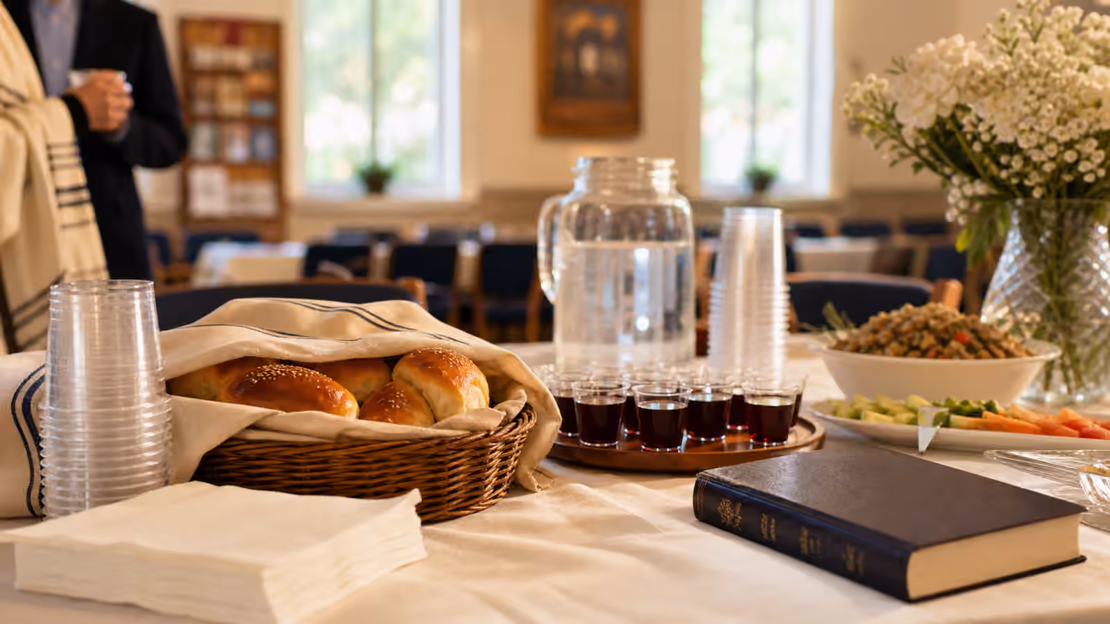 A synagogue social-room table with covered rolls, small cups, water, platters, flowers, and a closed prayer book.