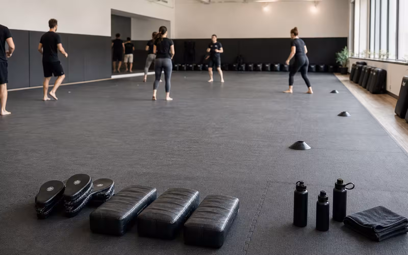 A calm Krav Maga training floor with pads, cones, water bottles, open space, and adults warming up under soft studio light