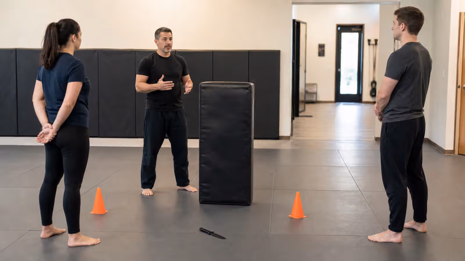 A Krav Maga instructor discussing a cautious weapon-awareness drill with a padded barrier, cones, and a rubber training prop.