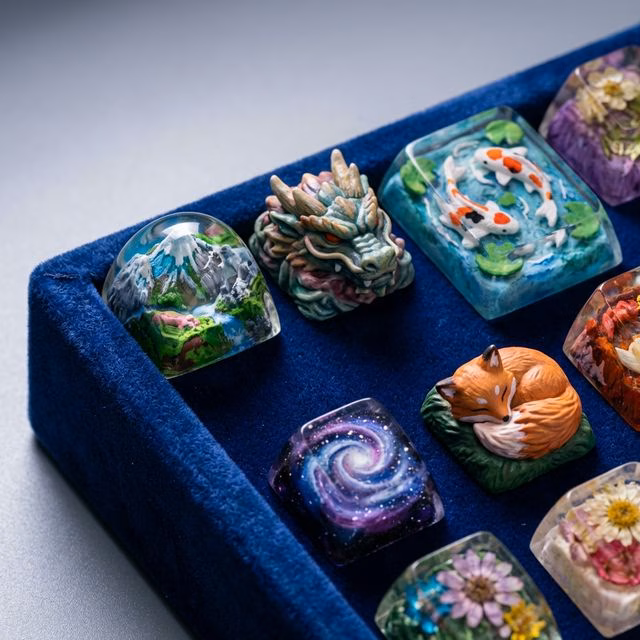 Collection of colorful artisan keycaps on a velvet display tray showing unique hand-crafted designs including miniature landscapes, sculpted creatures, and resin art pieces, soft studio lighting, realistic macro product photography