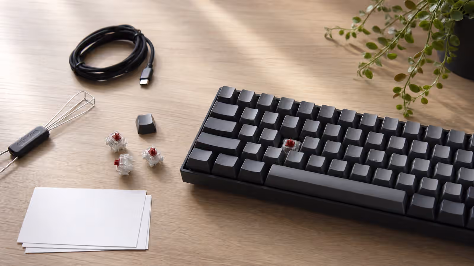 Compact mechanical keyboard with one keycap removed, loose switches, a puller, cable, and blank cards on a clean desk.