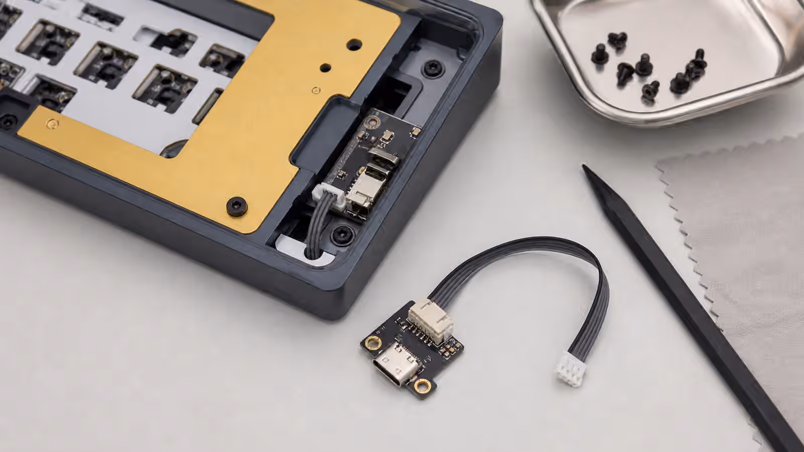 Opened keyboard case with USB-C daughterboard, small cable, screws, spudger, and cloth on a clean workbench.