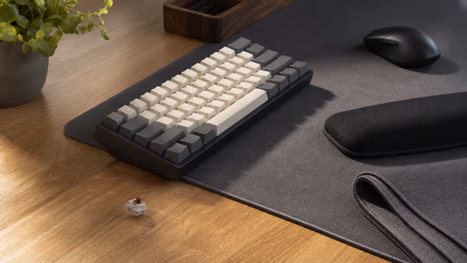 Compact mechanical keyboard on a thick desk mat beside a bare wood desk edge, mouse, wrist rest, and mat sample.