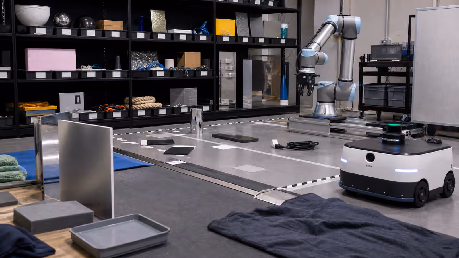 A robotics lab shelf and test lane with varied objects, reflective panels, cables, trays, a robot arm, and a mobile robot.