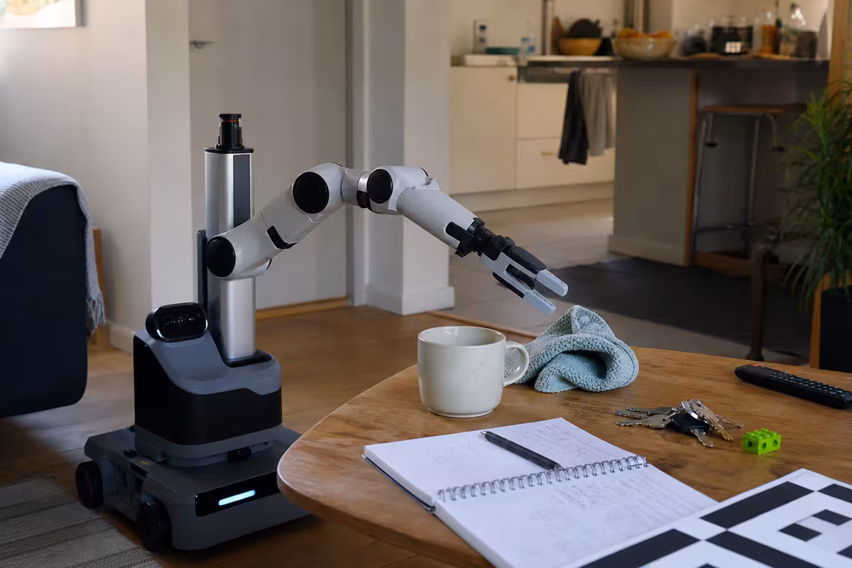 A compact home robot arm reaches toward a mug and dish towel on a low table with everyday household objects nearby.