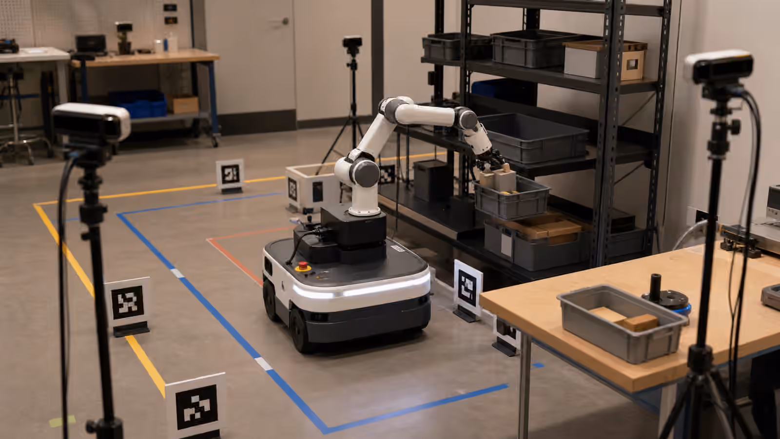 A mobile robot with an arm aligning beside shelves, bins, cameras, and a marked floor path in a robotics lab.
