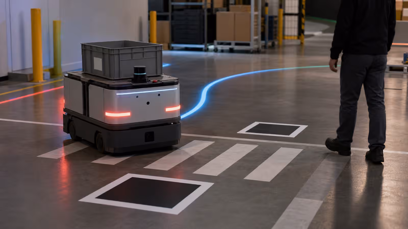 A mobile robot with status lights paused at a marked crossing while a worker stands at a safe distance.
