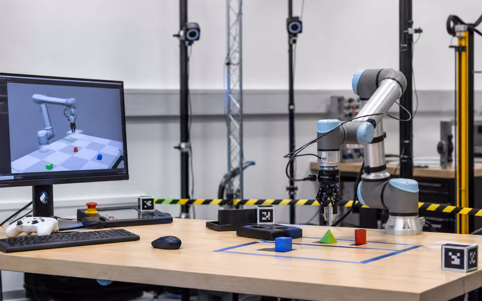 A robotics lab with a robot arm, test objects, motion-capture markers, safety tape, and a monitor showing an unreadable simulated robot scene.