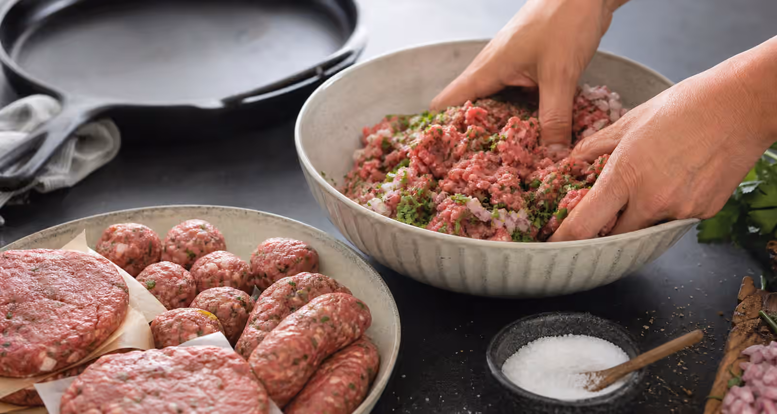 Ground meat being mixed with salt, herbs, and onion beside a cast iron skillet.