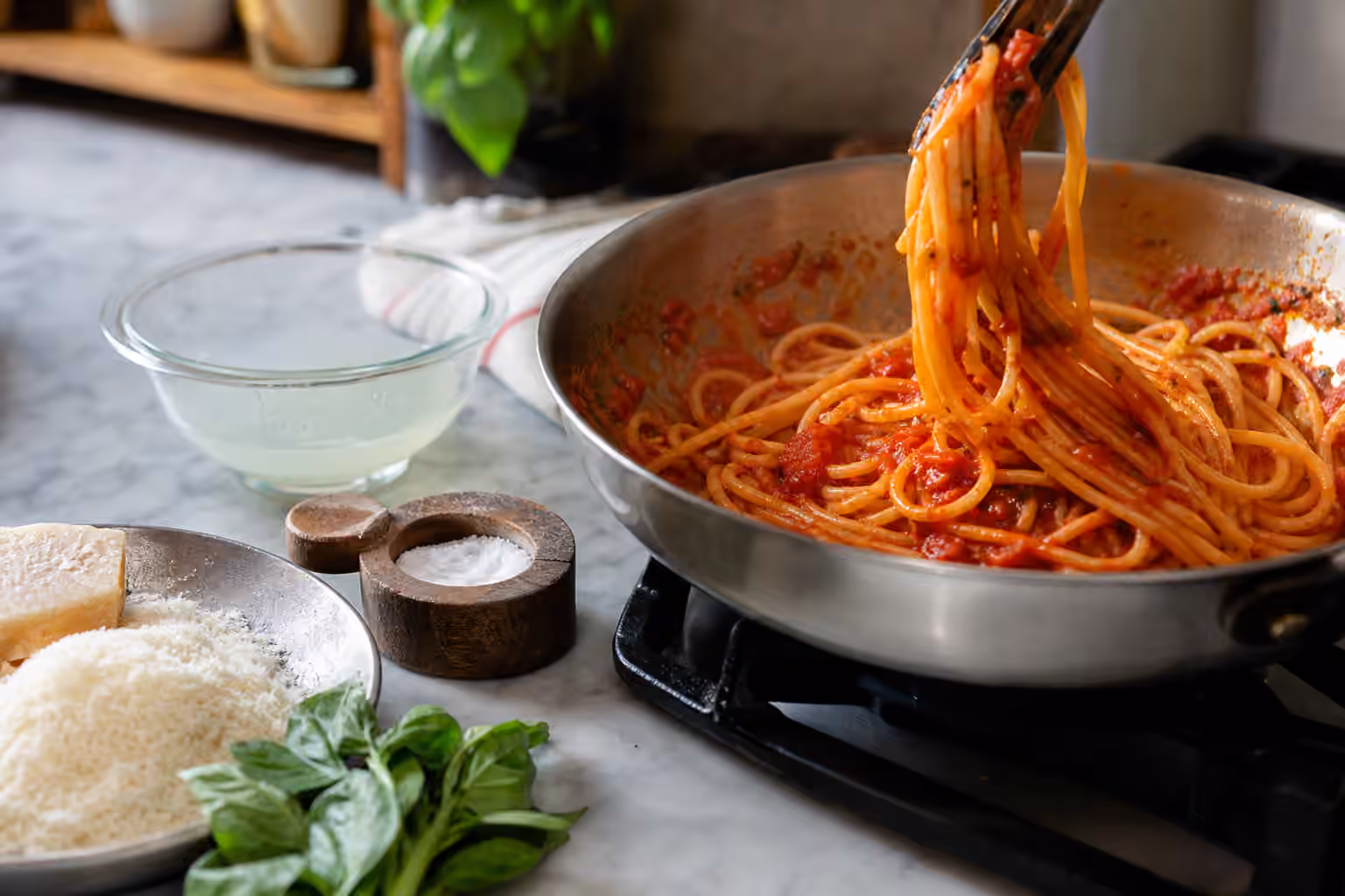 Pasta being tossed with tomato sauce beside pasta water, cheese, basil, and a salt cellar.