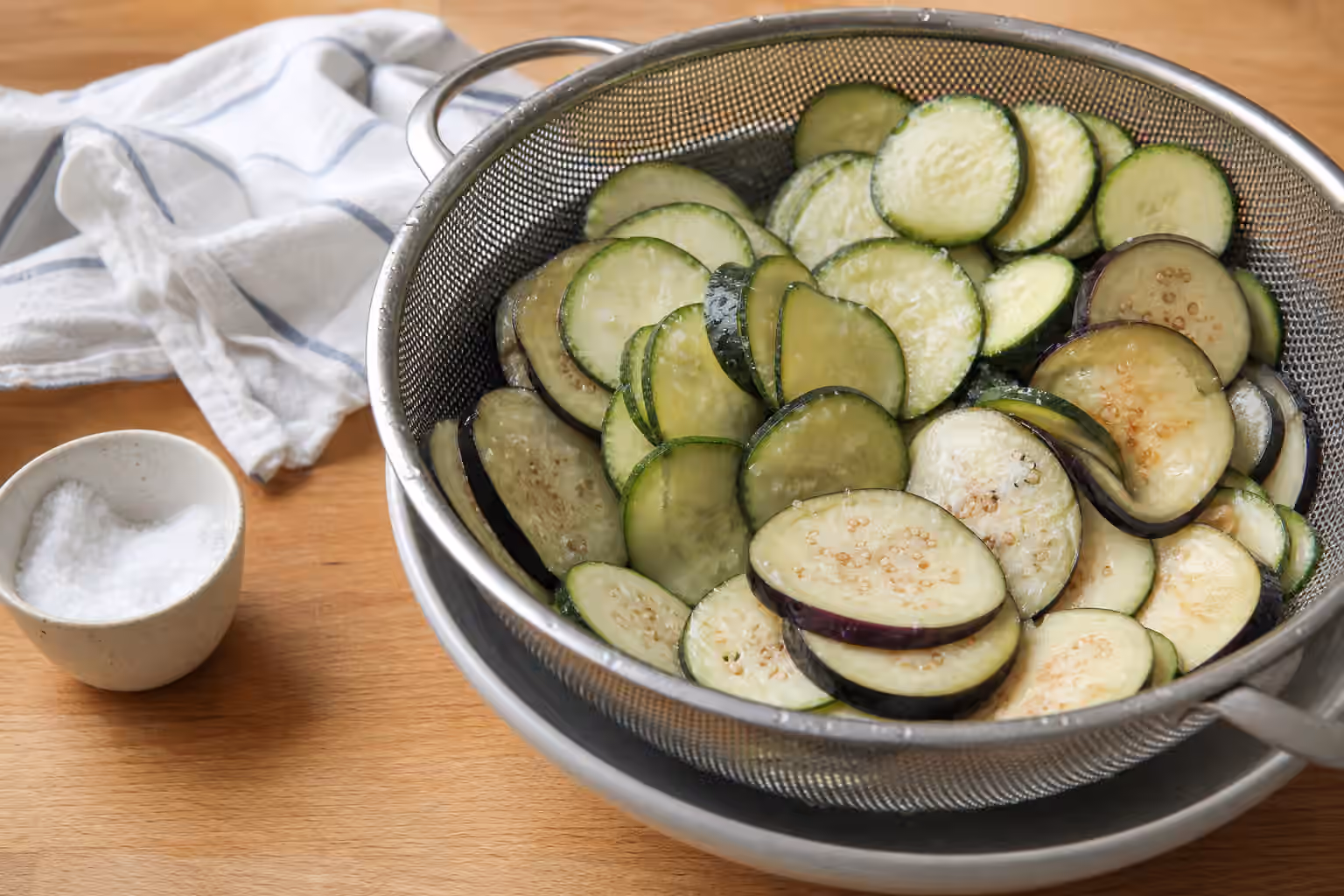 Sliced cucumbers, zucchini, and eggplant draining in a colander after salting.