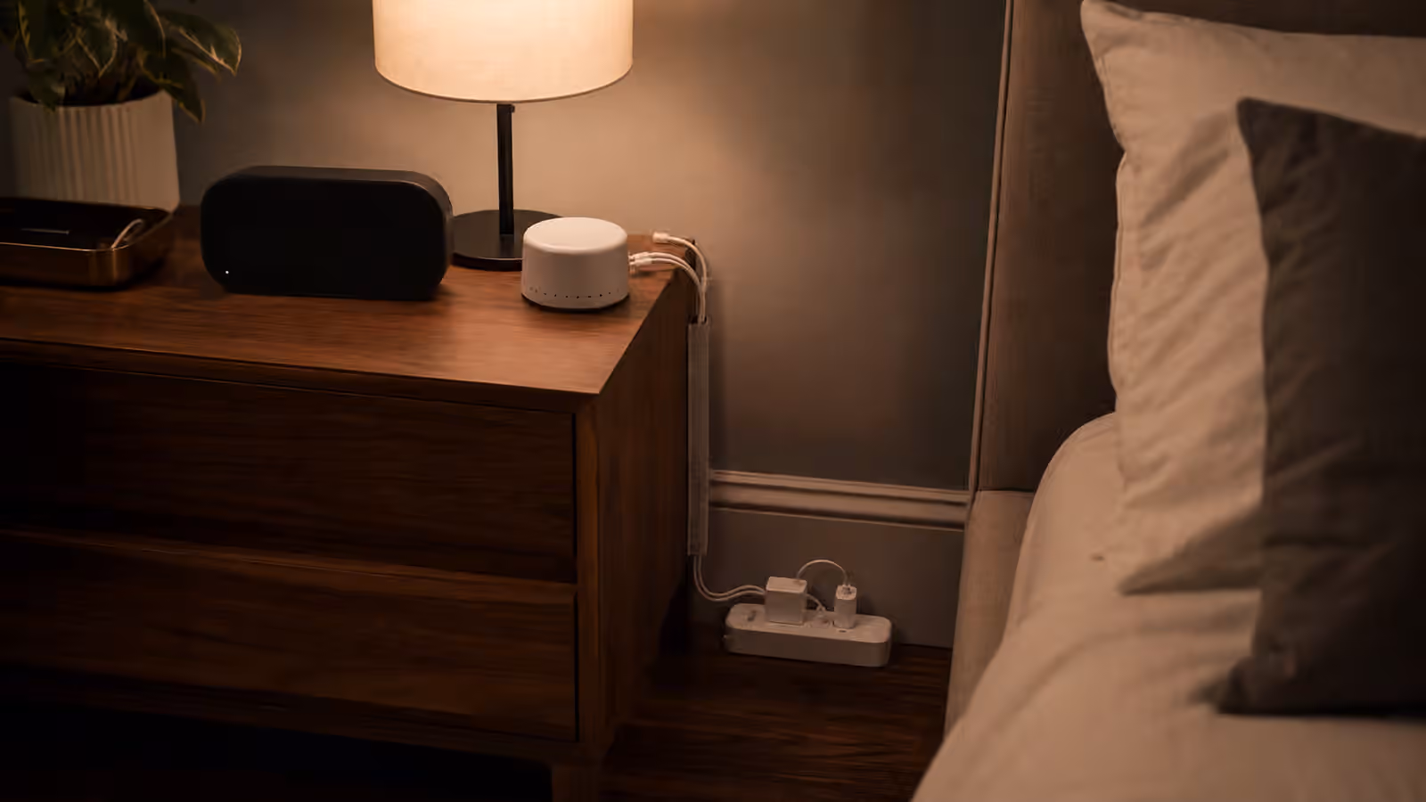 A quiet bedroom nightstand with dim electronics, routed charging cables, a small sound machine, and a warm lamp.
