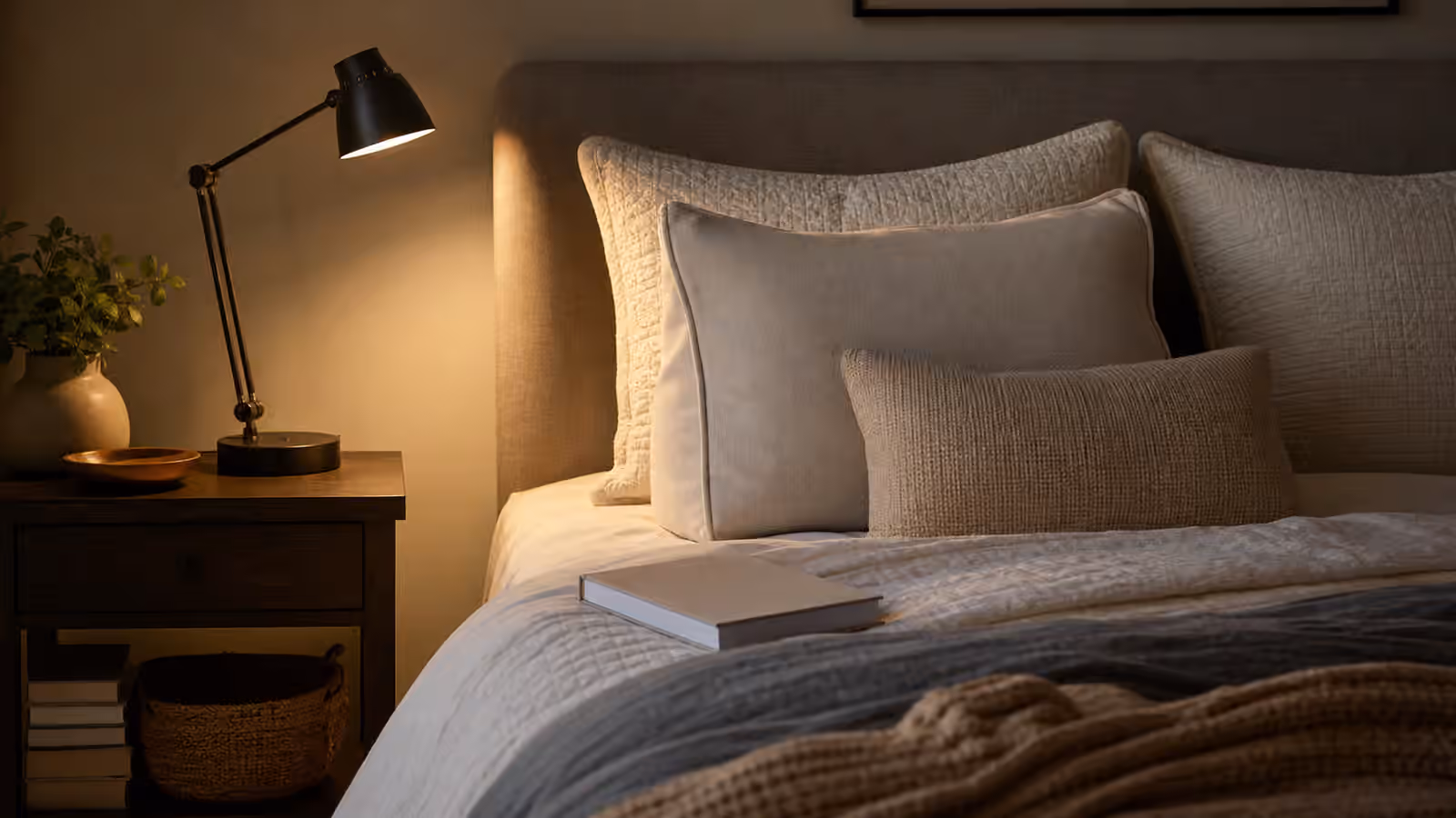 A warm bedside reading lamp beside layered pillows, a book, and a tidy nightstand in a calm bedroom.