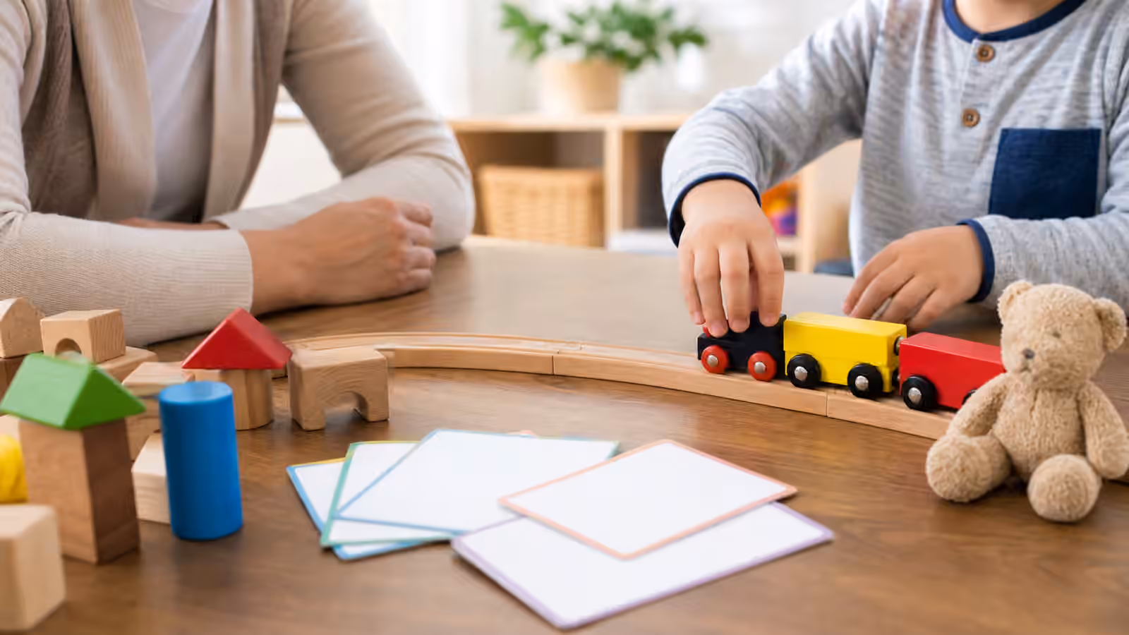 Preschool play table with a toy train, wooden blocks, blank cards, a stuffed animal, and adult and child hands.