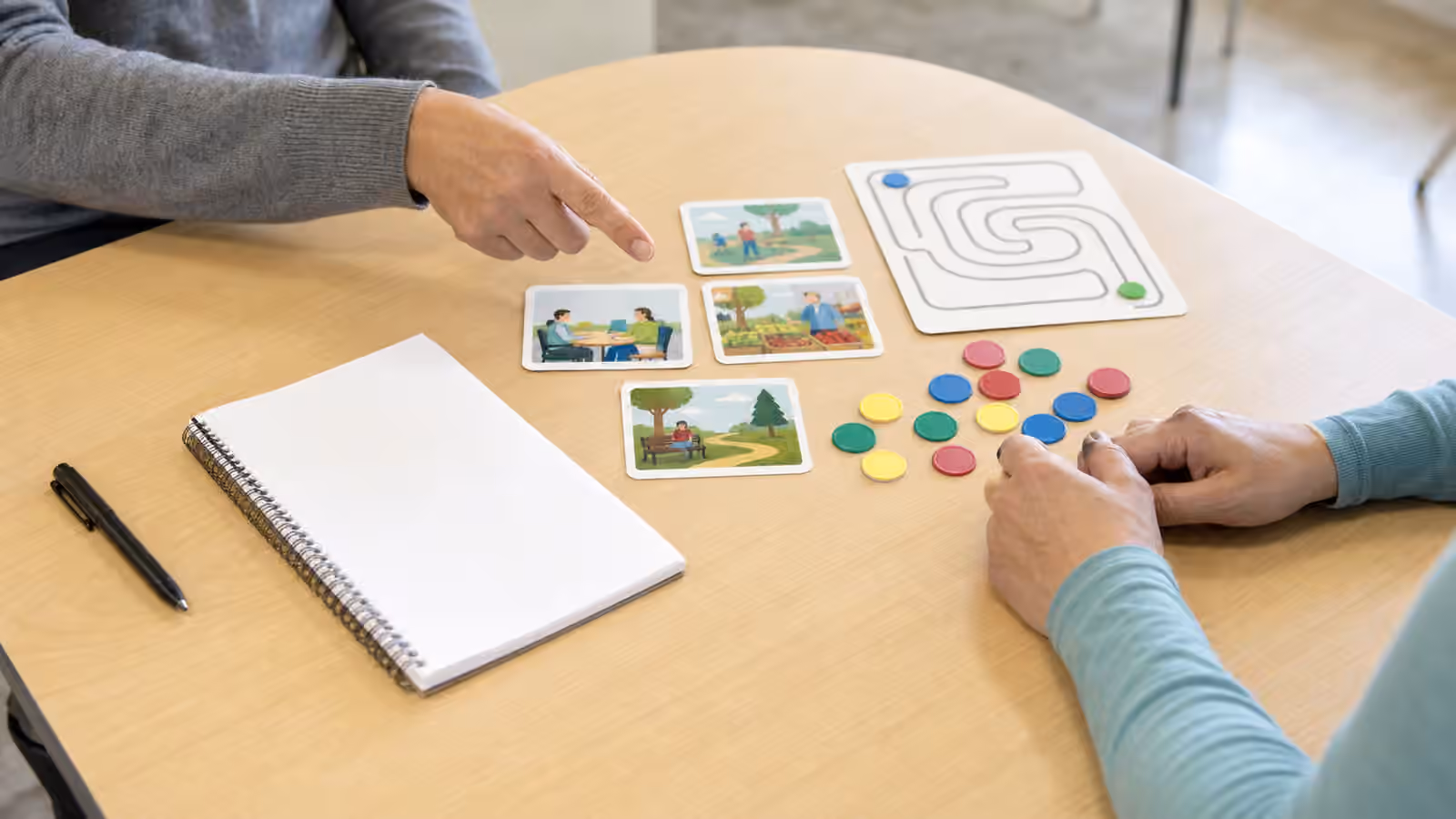 Rehabilitation table with a blank notebook, picture cards, route card, colored tokens, and two adults' hands.