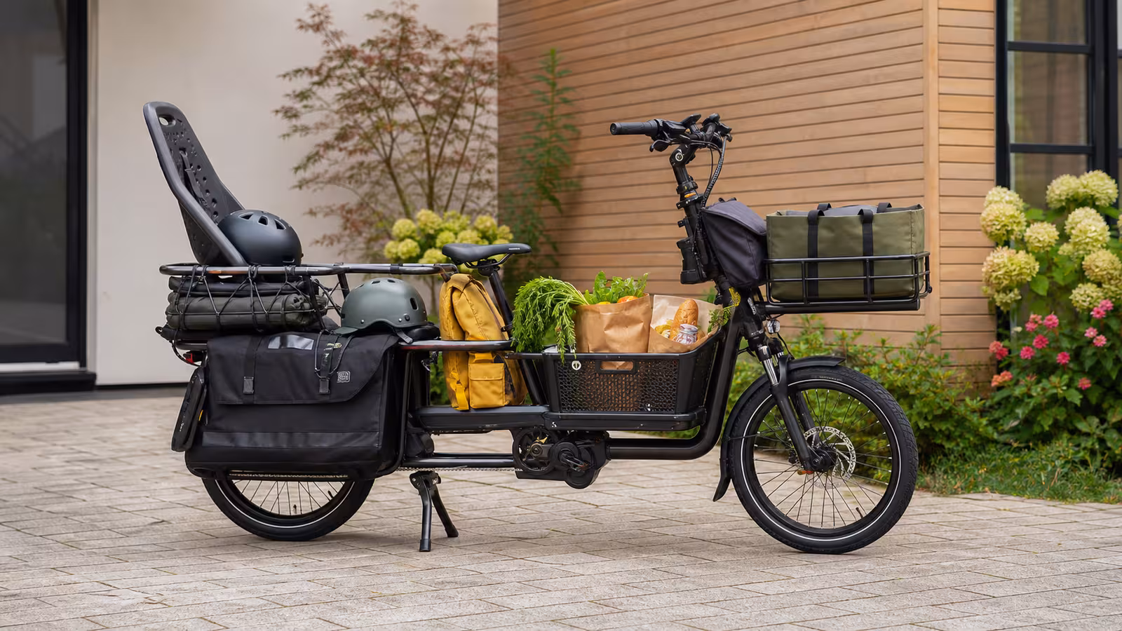 A longtail electric cargo bike parked in a courtyard with panniers, a front basket, groceries, a child seat, helmets, a school backpack, and a folded rain cover.