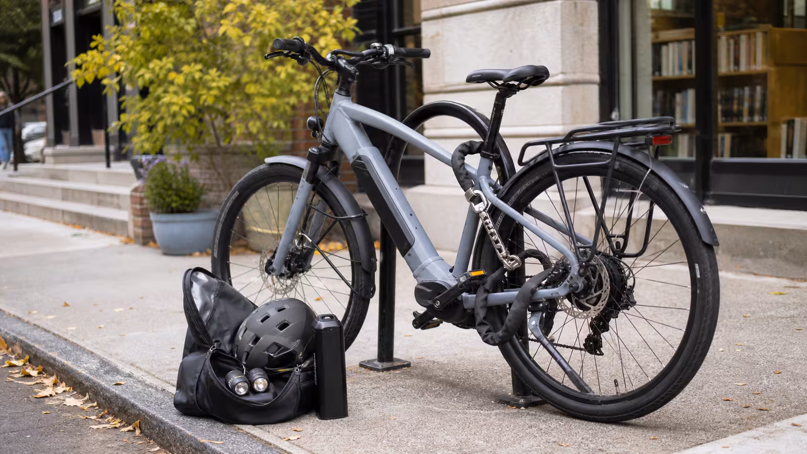 An electric bike locked to a sturdy city rack with a U-lock and chain near a bag holding removable lights and a helmet.