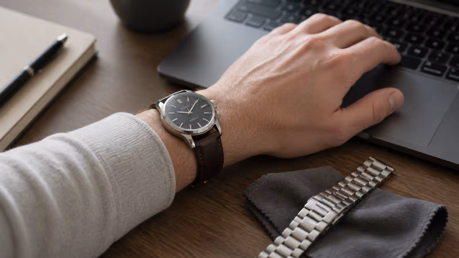 A wrist wearing an unbranded watch rests near a laptop keyboard with a spare bracelet and cloth nearby.