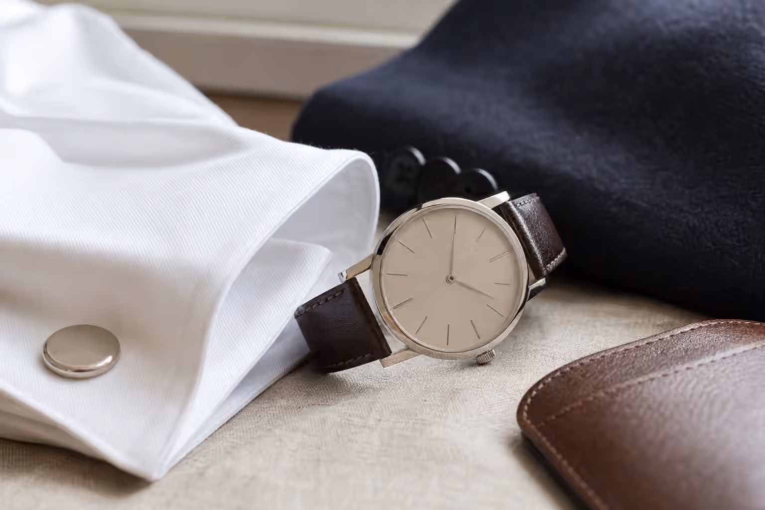 A slim unbranded dress watch rests beside a white shirt cuff, dark jacket sleeve, and brown leather strap on a tailor's table.